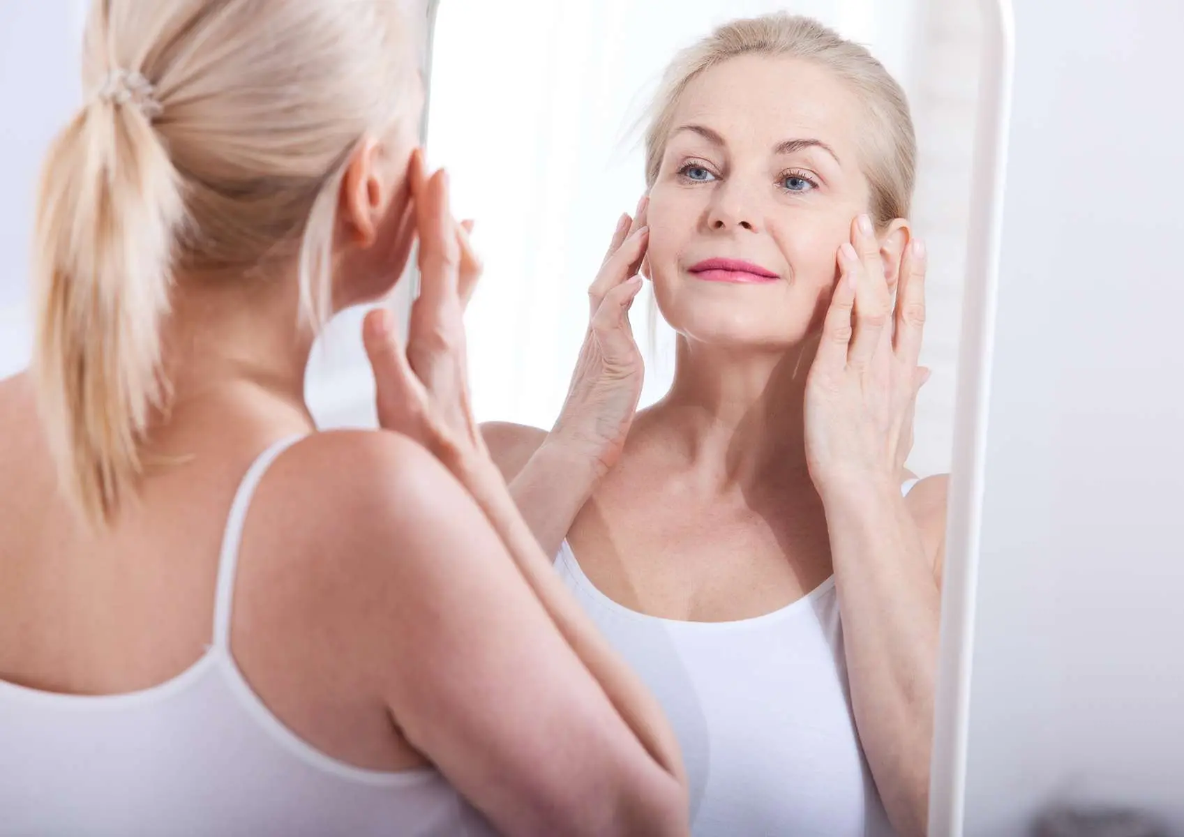 Femme senior avec une belle peau qui se touche les joues avec les mains devant un miroir