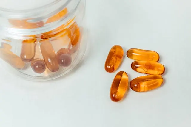 Bocal transparent avec capsules compléments alimentaires oranges et  5 capsules oranges à côié sur fond vert d'eau